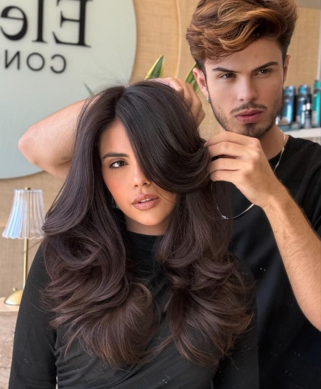 Lucas Barboza finalizando um corte em cabelo longo e escuro, com o painel do salão ao fundo
